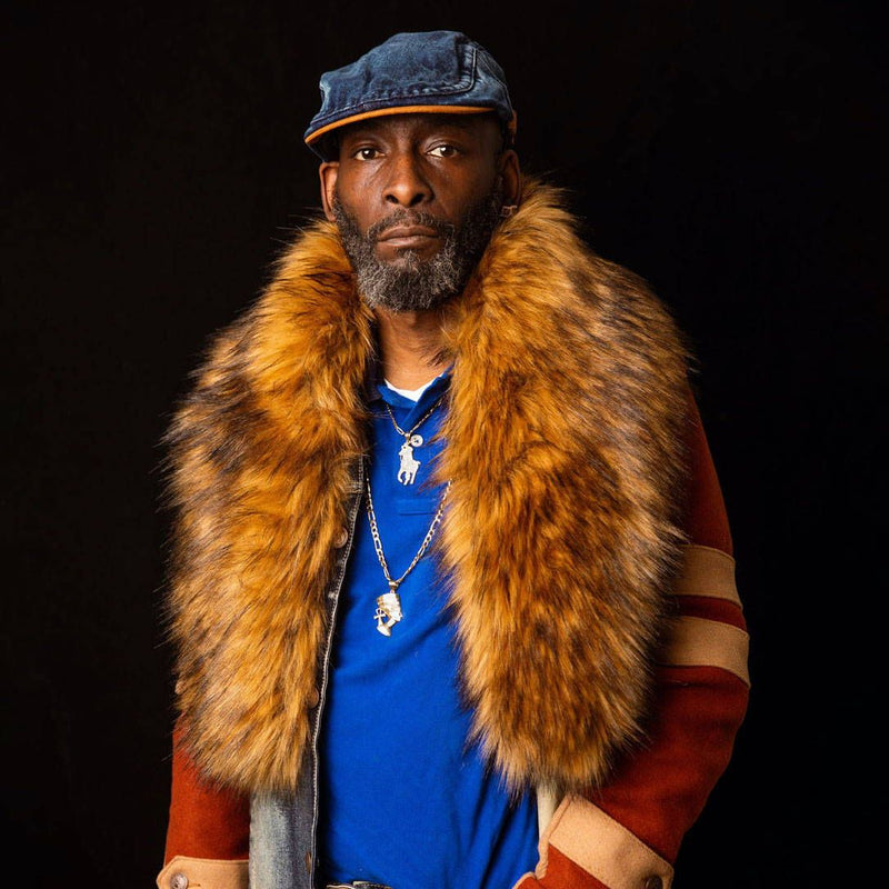 Tony stares at the camera wearing a bright blue polo and fur shawl against a black background.