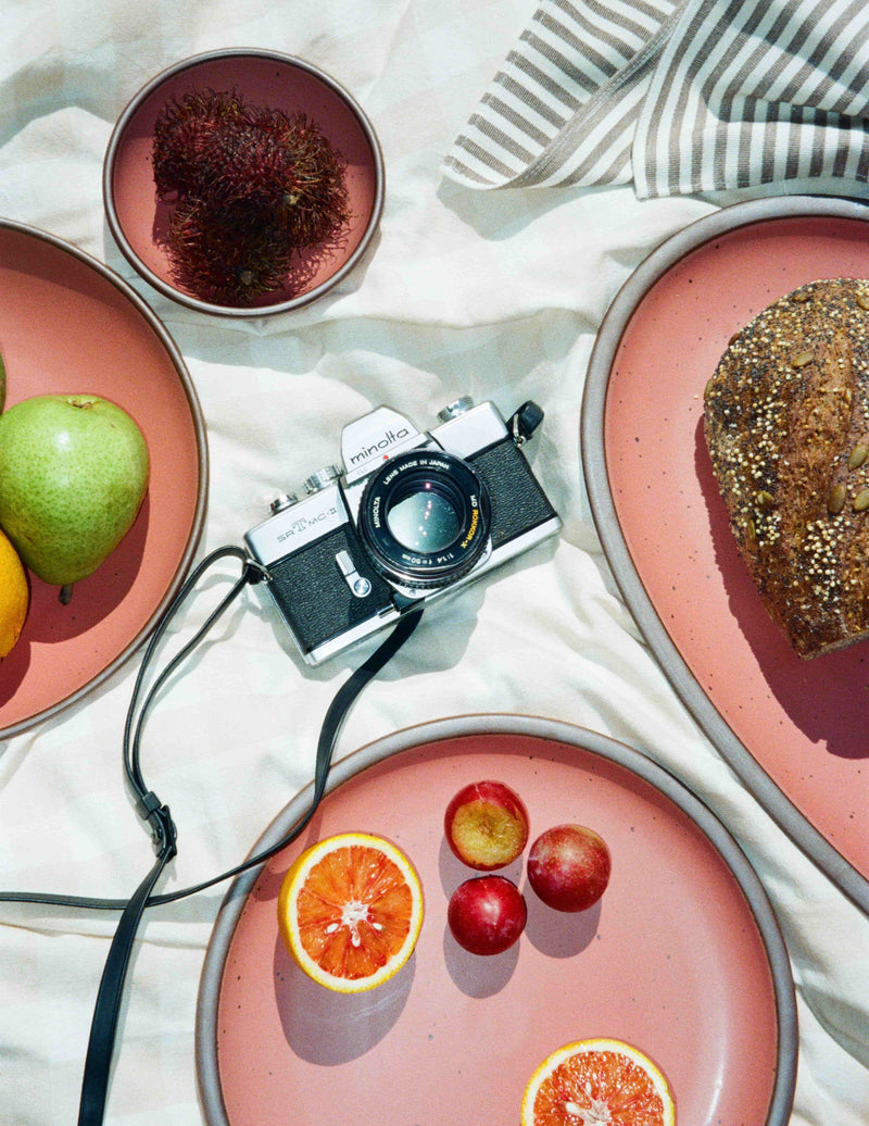 Minolta film camera surrounded by fruit and bread on watermelon pink ceramic plates over a white picnic cloth.