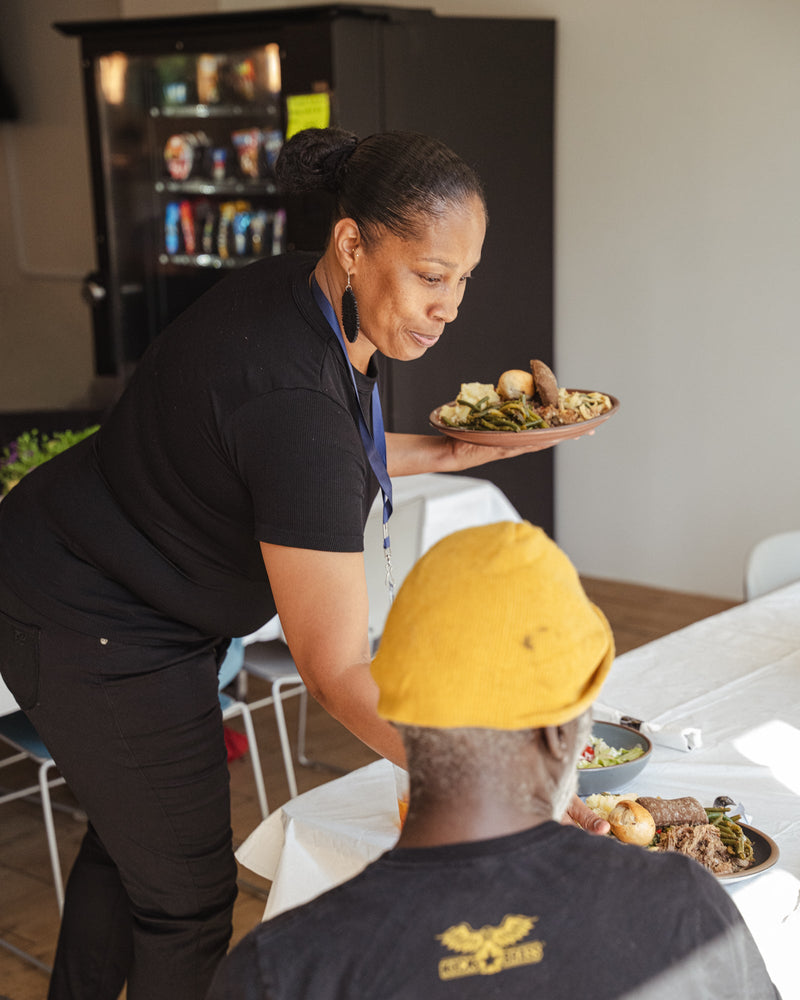 A woman sets a plate of food down on a table in front of a seated person