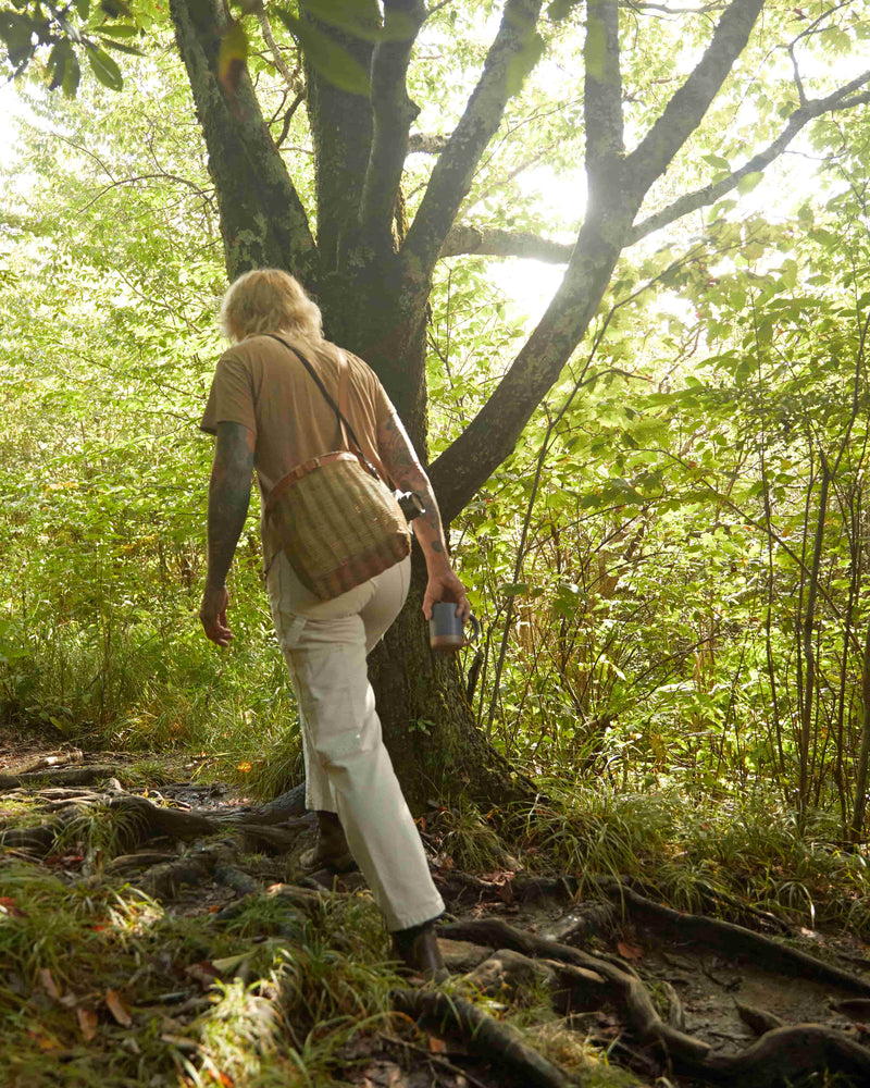 A person walking away in the woods holding a basket bag and carrying a blue mug