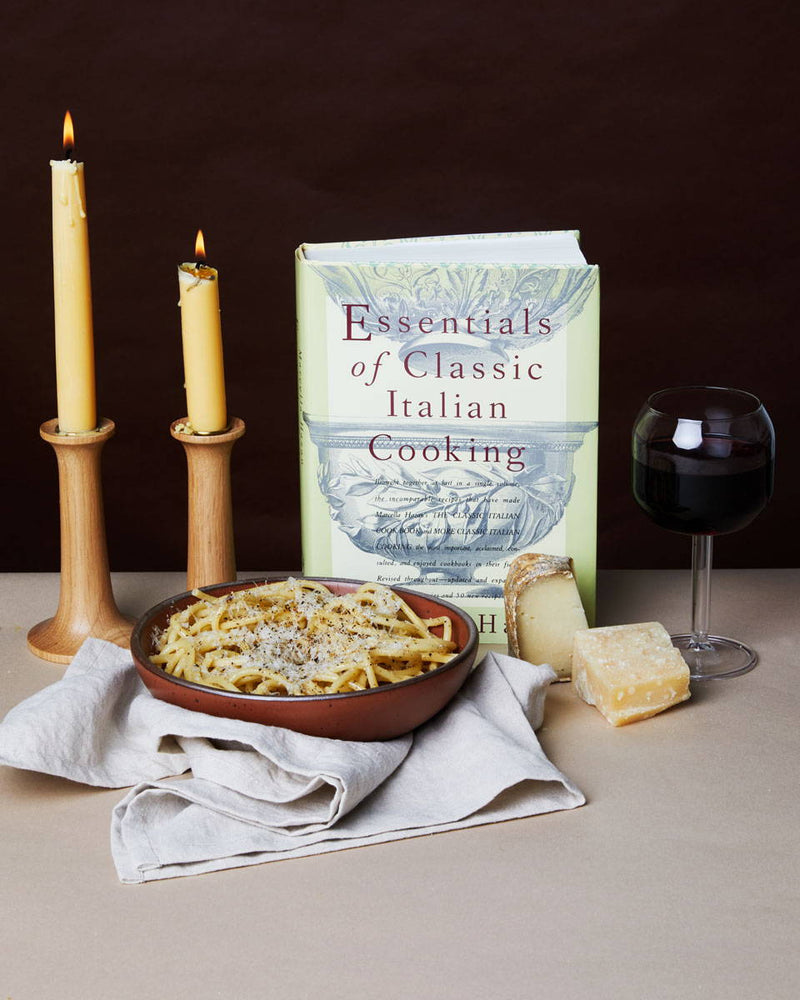 An Italian Cookbook is propped up and surrounded by a bowl of pasta, whole parmesan cheese, a glass of red wine, and lit taper candles