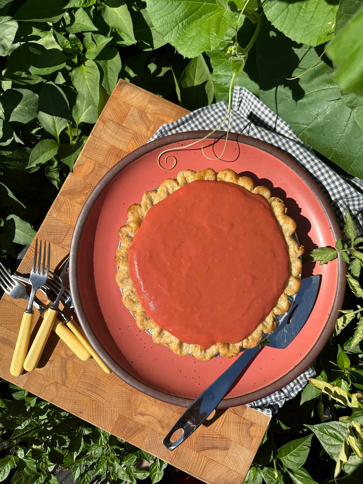 A golden-brown pie topped with smooth guava filling sits on a watermelon pink plate with brown rim, surrounded by greenery and utensils.