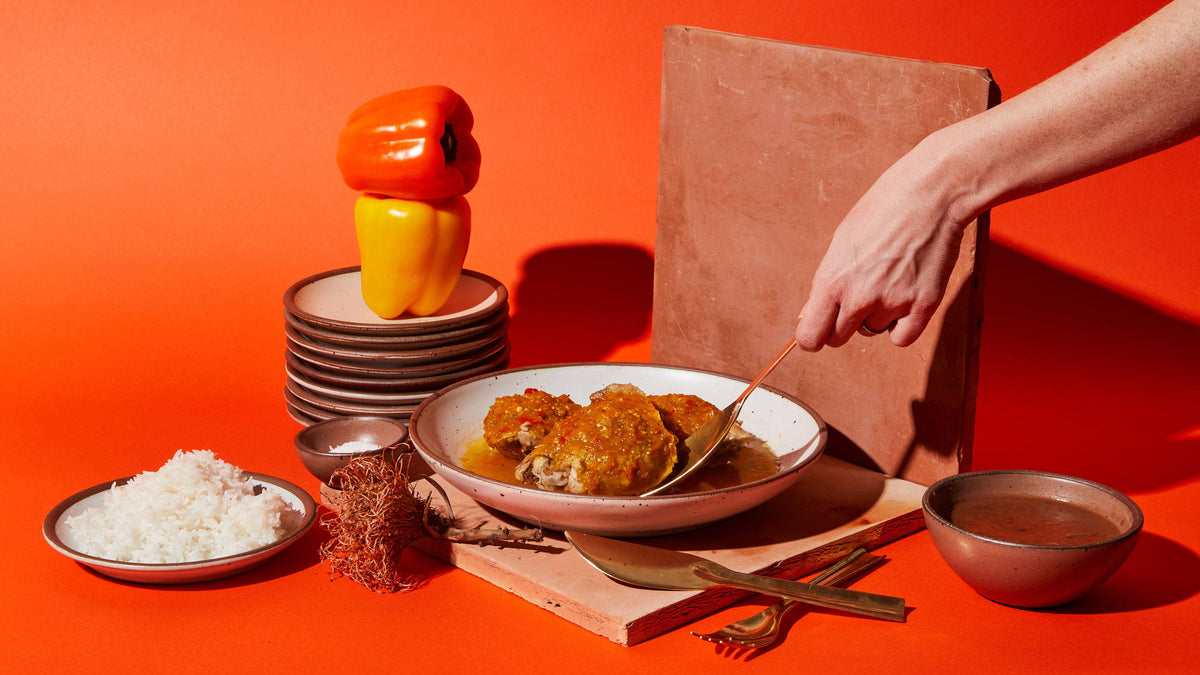An artful layout of a white coupe bowl with an entree and a hand reaching to grab a piece with a brass spoon. Surrounding are a plate of rice, bell peppers on a stack of plates, and more.