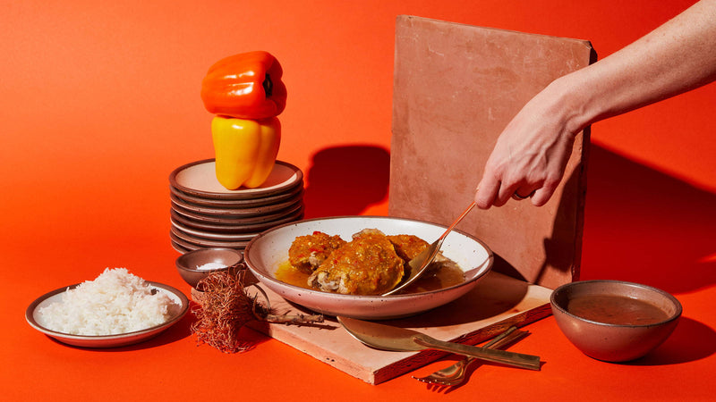 An artful layout of a white coupe bowl with an entree and a hand reaching to grab a piece with a brass spoon. Surrounding are a plate of rice, bell peppers on a stack of plates, and more.