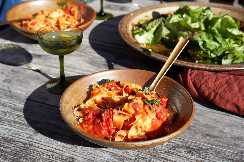 Shallow bowls of pasta and a large bowl of salad with a green coupe glass on an outdoor table.