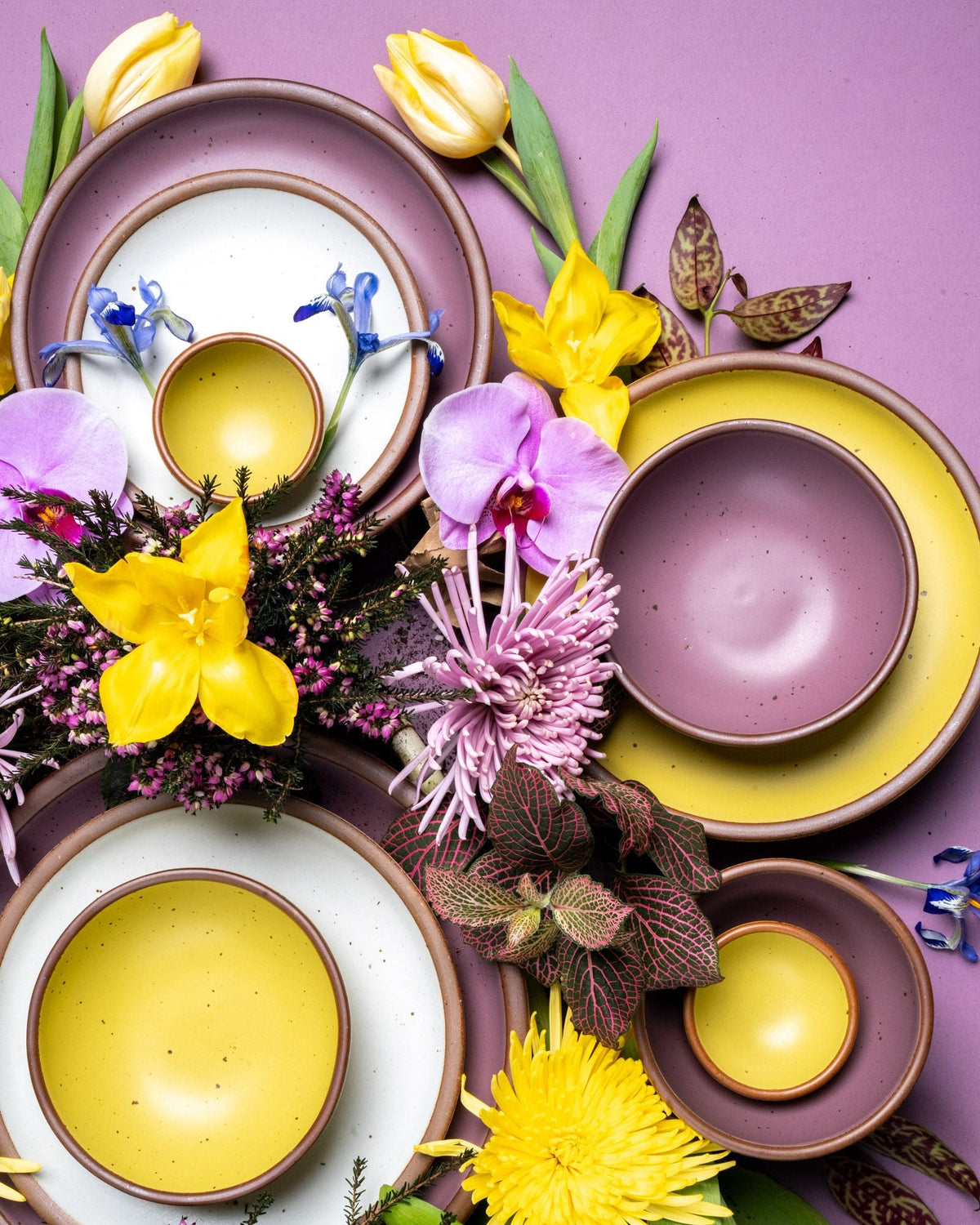An overhead view of colorful ceramic bowls and plates with bright flowers and leaves, all in yellow, purple, and pink colors.