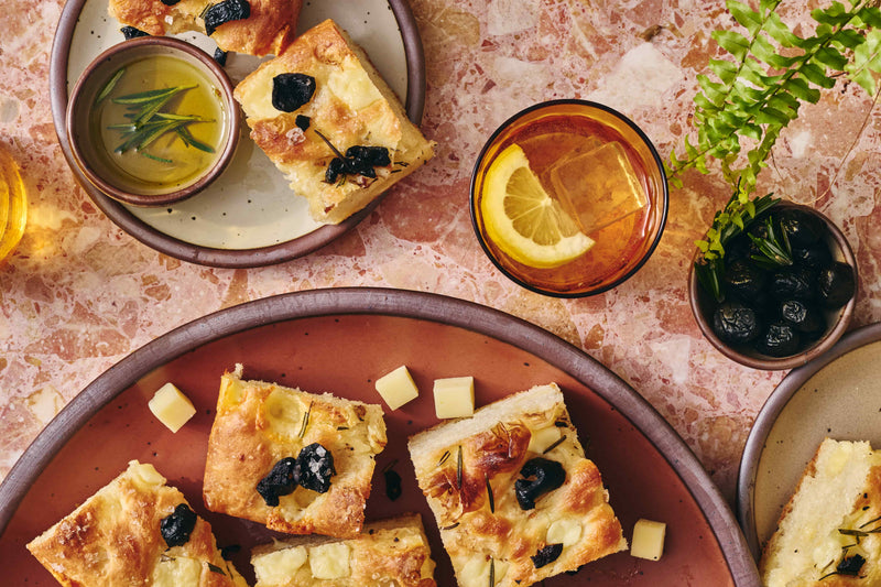 Slices of focaccia bread on a large ceramic plate, and smaller plate, artfully arranged next to Cabot Creamery cheese cubes, and a glass filled with water and lemon.