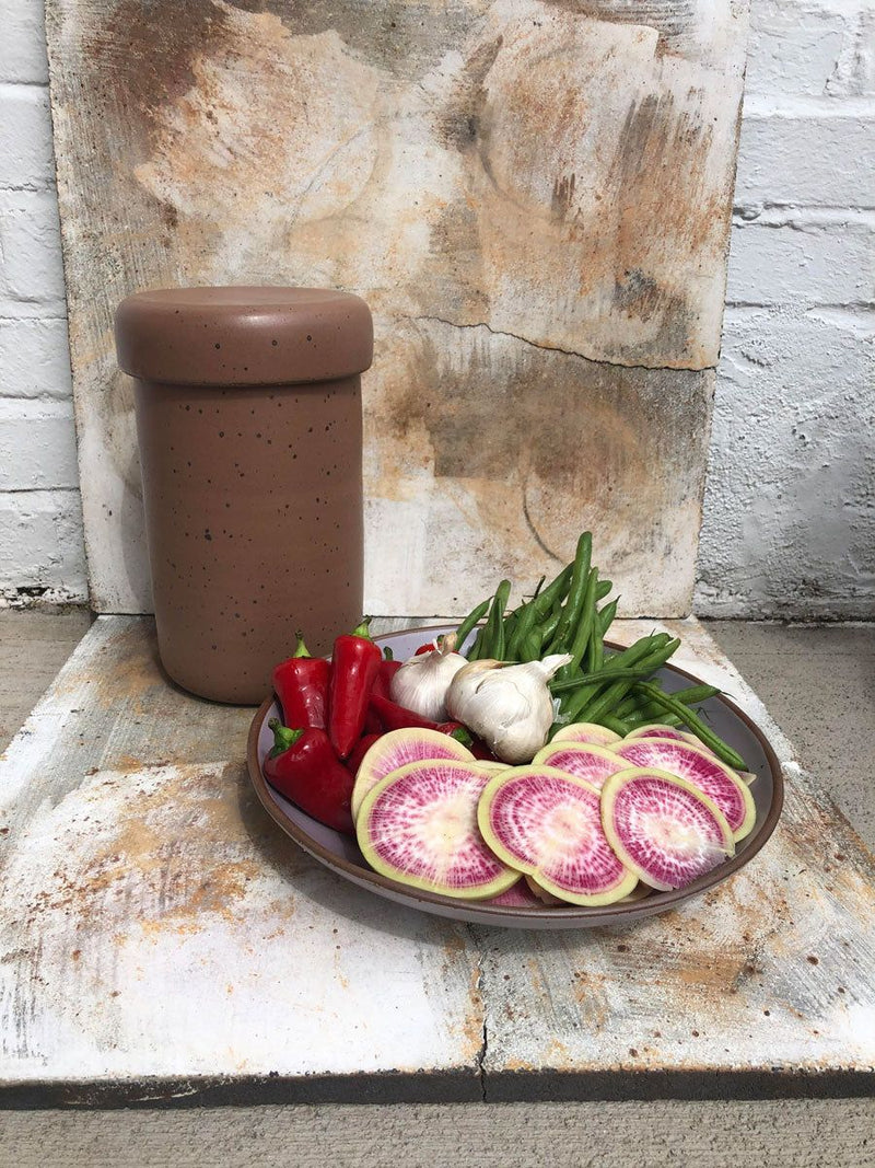A plate of raw vegetables to be fermented next to a ceramic crock with a lid.