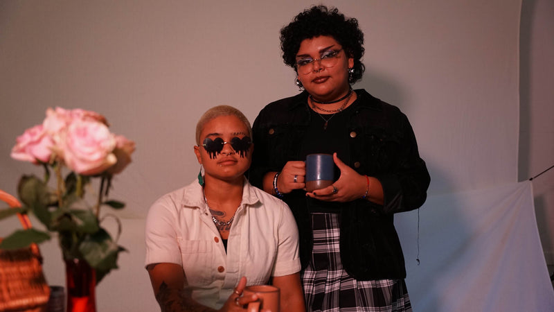 A wide frame shot of two people, one with short hair and melting heart sunglasses sitting down and the other with glasses and dark curly hair standing up holding a mug