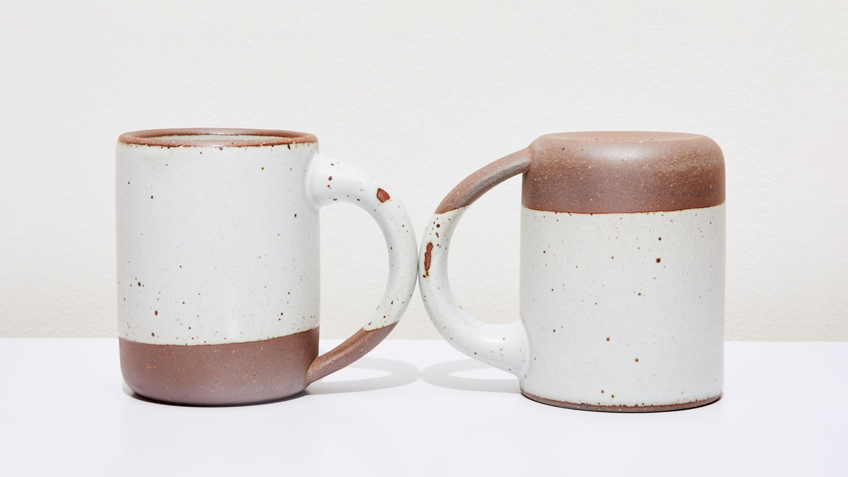 Two white ceramic mugs, one sitting up right, one upside down, with little glaze chips on the handles.
