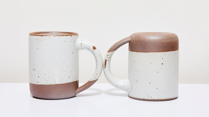 Two white ceramic mugs, one sitting up right, one upside down, with little glaze chips on the handles.