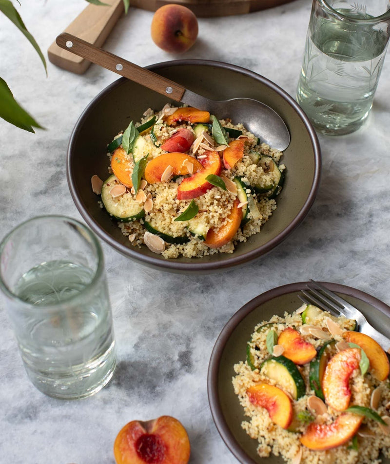 Two olive green bowls sit on a marble countertop filled with couscous and sliced peaches and surrounded by glassware and peaches.