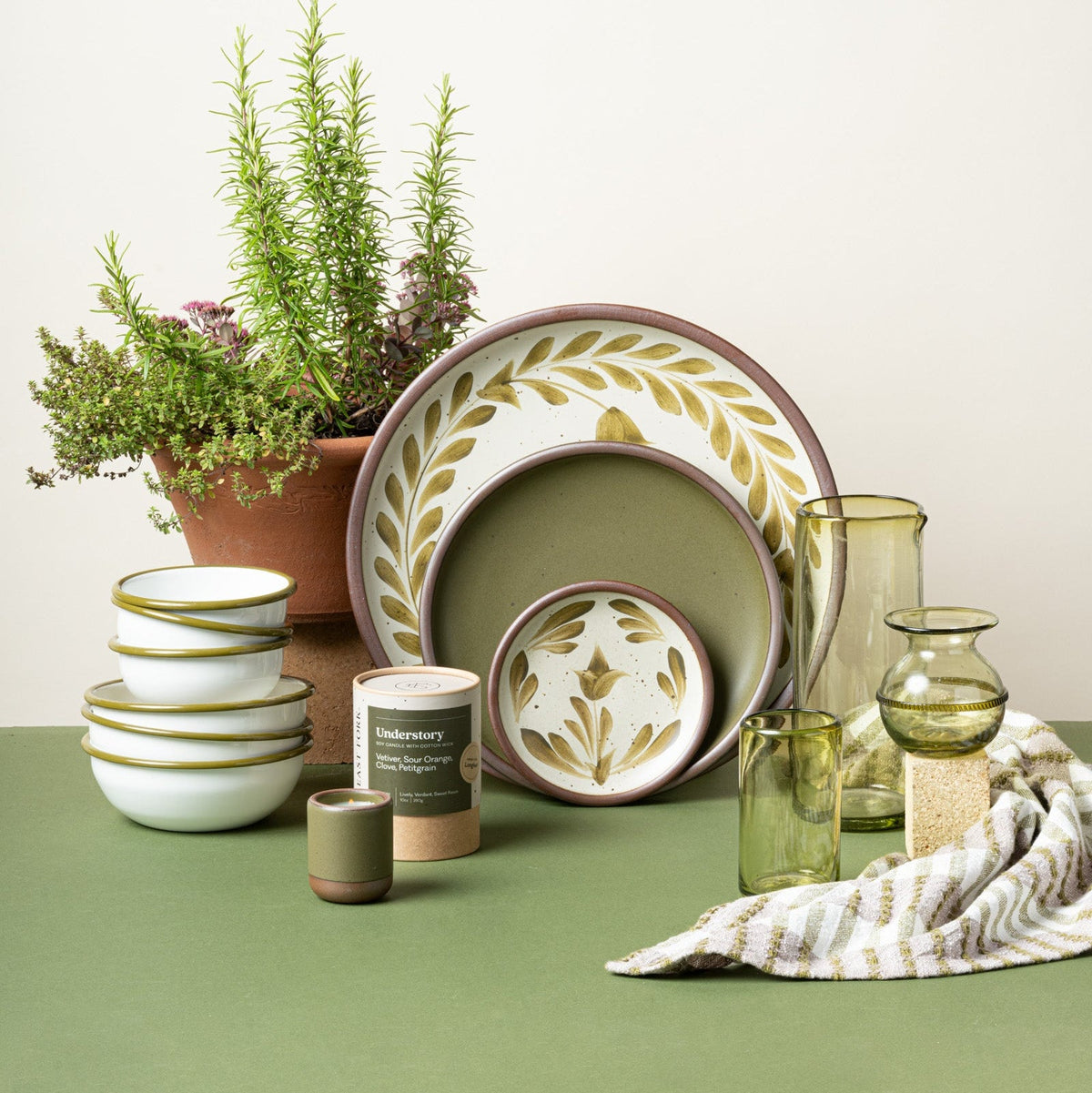 Styled tabletop with hand-painted green pottery, stacked enamel bowls, green glassware, an Understory candle, and potted herbs.