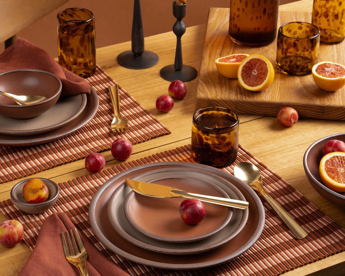 Rust-toned table setting with stoneware plates, amber glasses, plums, and blood oranges on striped placemats and a wood table.