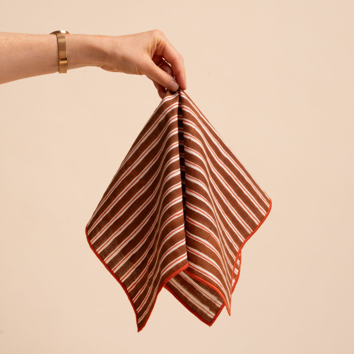 A hand holds out a striped cloth napkin with vertical brown and white stripes with orange accents and a stitched orange edge.