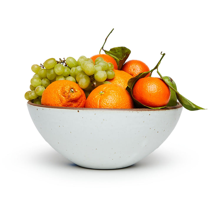 A large ceramic mixing bowl in a cool white color featuring iron speckles and an unglazed rim filled with a medley of fruit