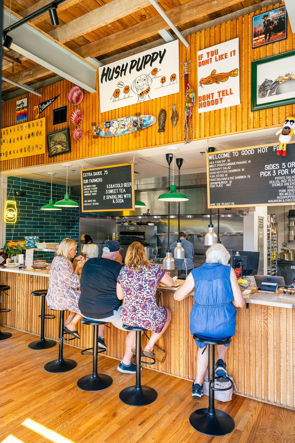 A counter at a restaurant kitchen is filled with people with colorful artwork and a menu above them