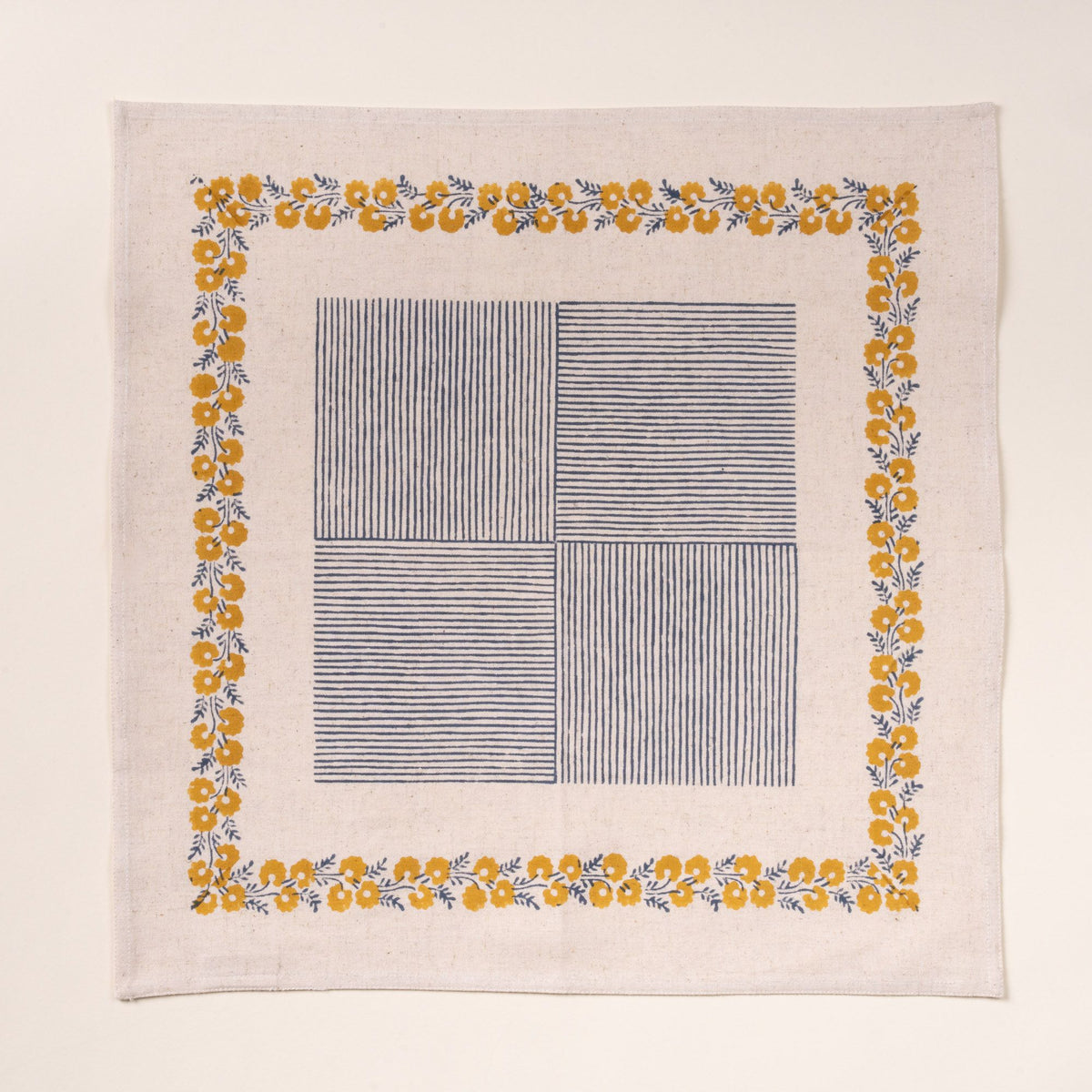 A cream-colored napkin with a delicate yellow floral border framing a central blue striped patchwork design, laid flat against a neutral background.