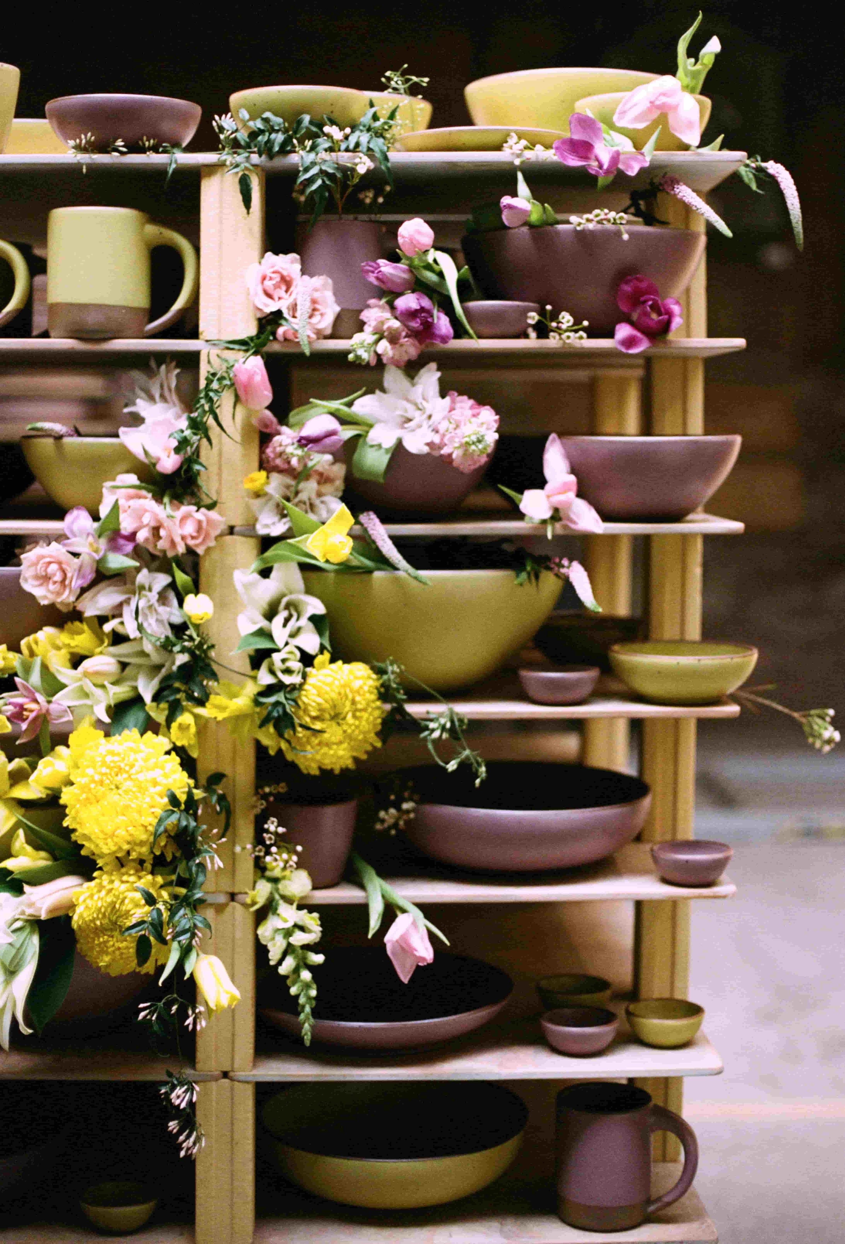 A photo of ceramic bowls, plates and mugs in Yuzu and Prune on a kiln cart in the factory, with adorning flowers in the same colors.