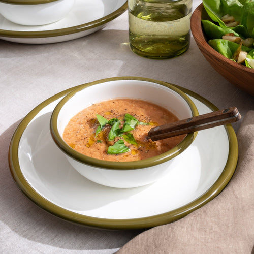 A small enamel bowl filled with tomato soup on an enamel plate, both dinnerware pieces are white with olive green rims.