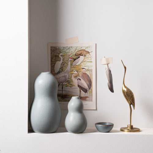 Light blue curvy vases and a matching bowl styled on a white shelf with a brass heron statue, bird illustration, and feathers taped to the wall behind.