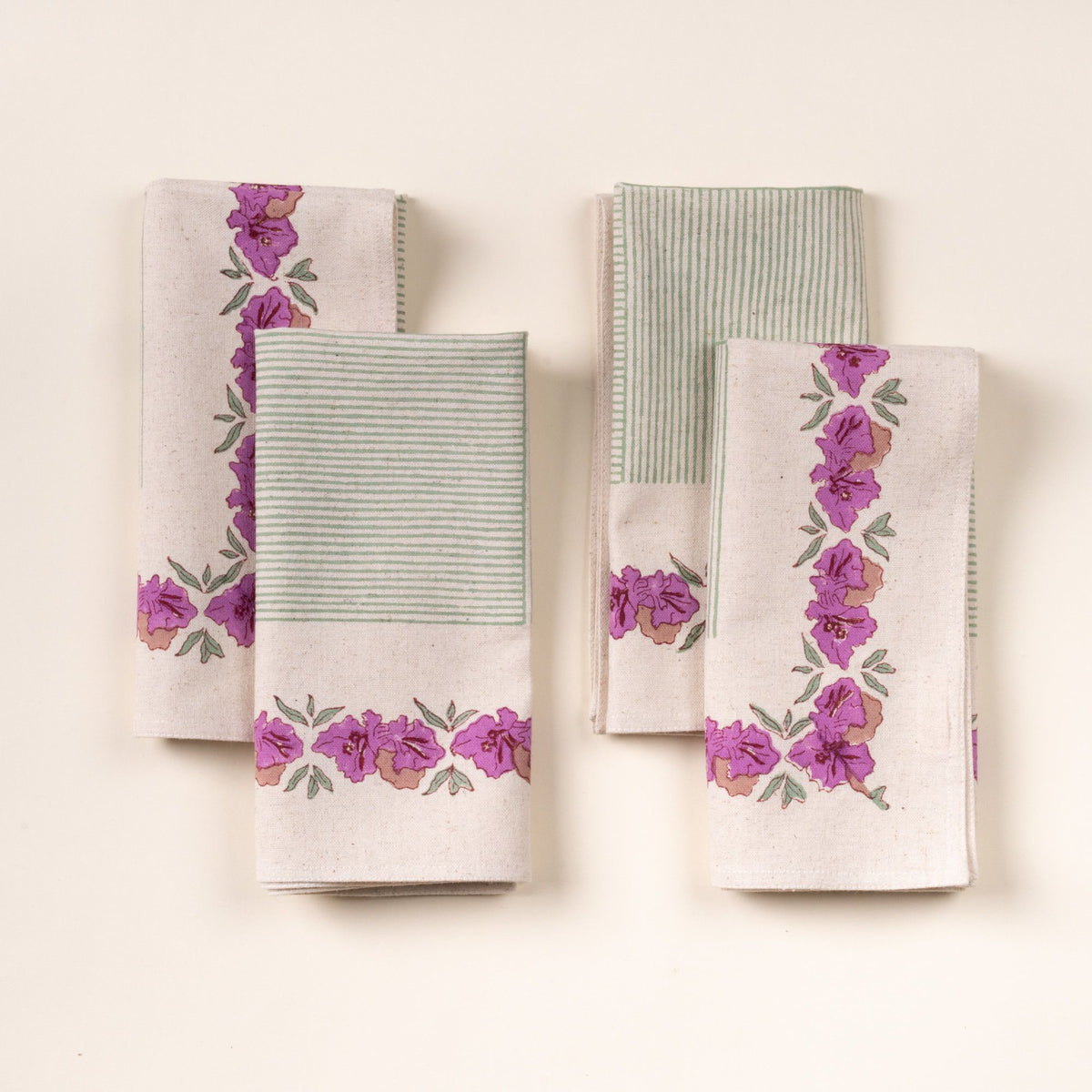 Four folded dinner napkins with green stripes and purple floral borders, arranged in two stacks on a cream background.
