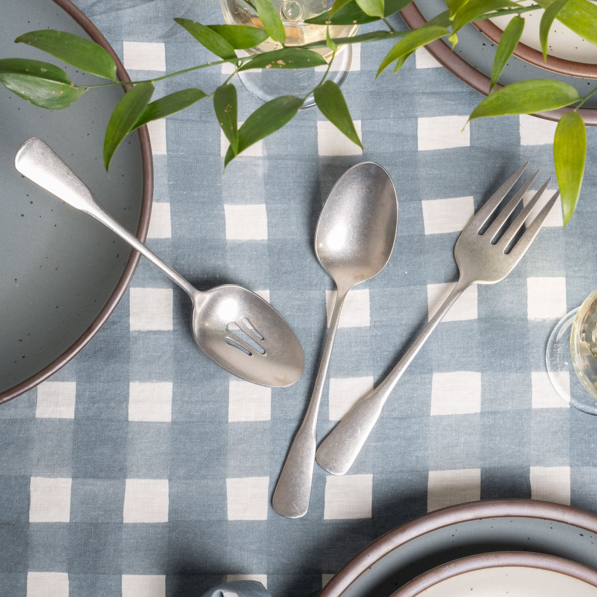 Three stainless steel serving utensils including a serving fork, serving spoon, and pierced spoon in a tumbled matte finish with a classic design, sitting on a blue-grey crosshatch tablecloth