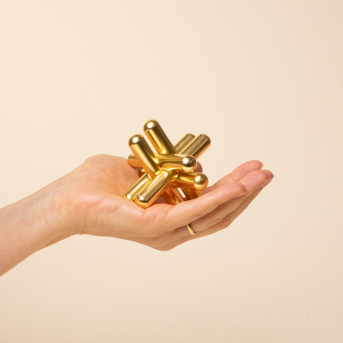 A hand holding a shiny gold interlocking metal puzzle sculpture with smooth cylindrical pieces on a beige background.