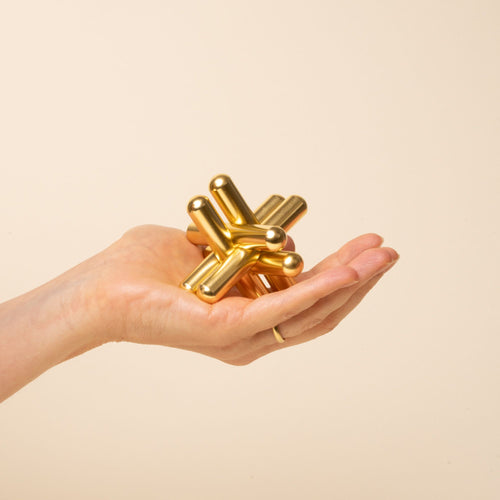 A hand holding a shiny gold interlocking metal puzzle sculpture with smooth cylindrical pieces on a beige background.