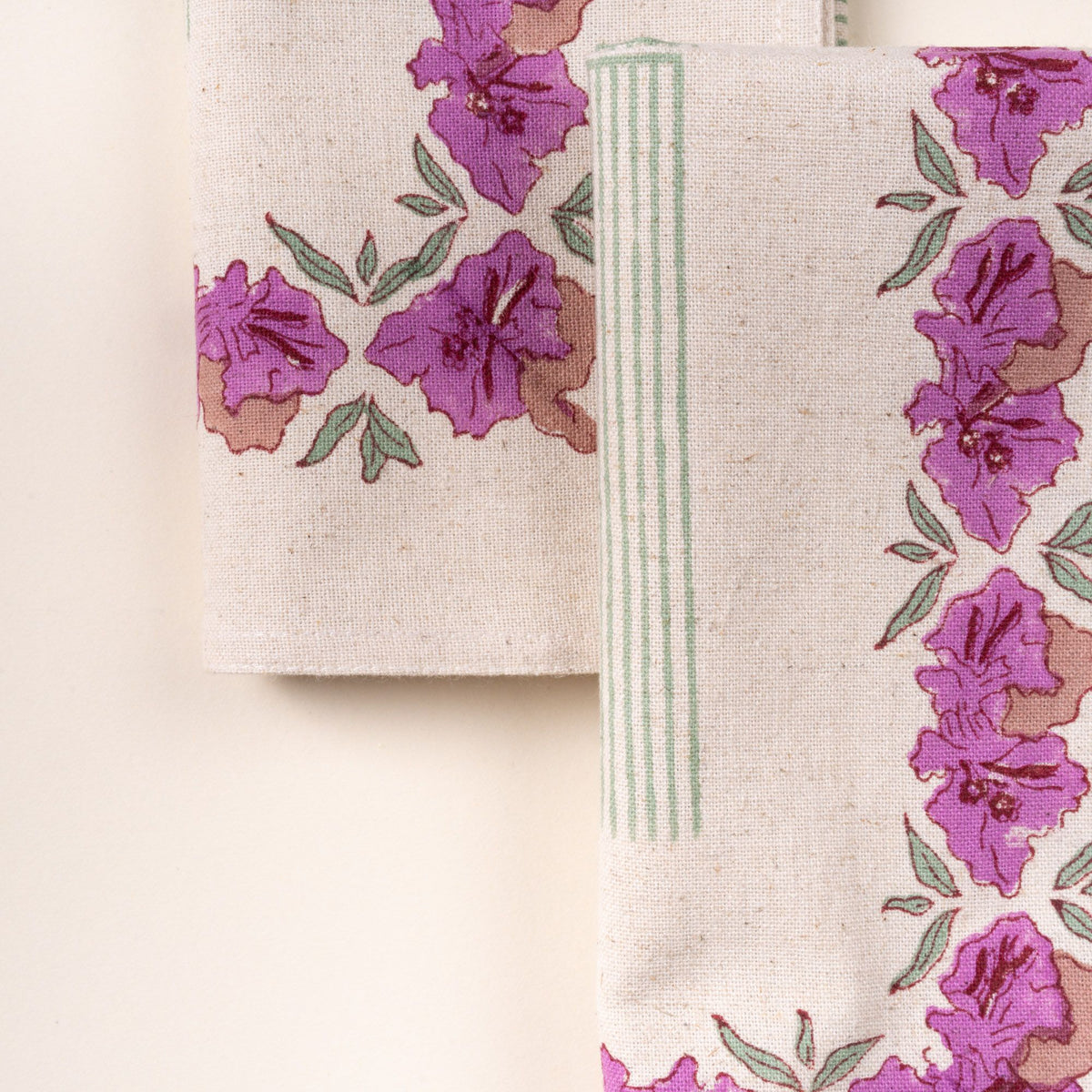 Close-up of folded linen napkins with purple floral prints and green striped accents on a cream background.