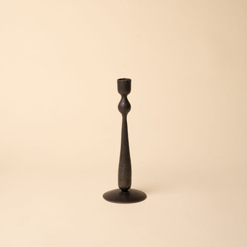 Tall minimal black forged iron candlestick with a sculptural shape on a neutral beige background.