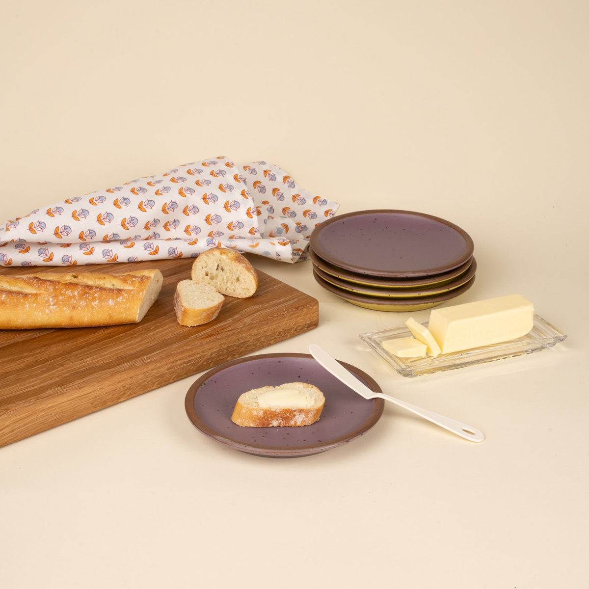 A white enamel butter knife sitting on a plate with a piece of bread and butter, and related items all around.