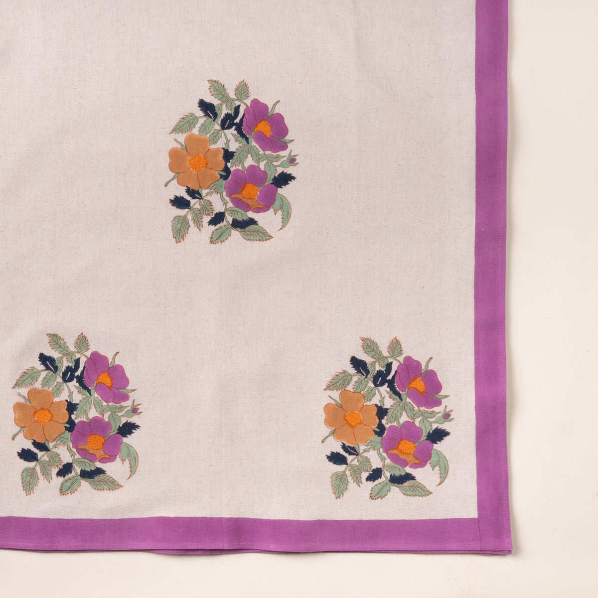 Cream linen tablecloth with a purple border and three floral motifs in purple and orange with green leaves.