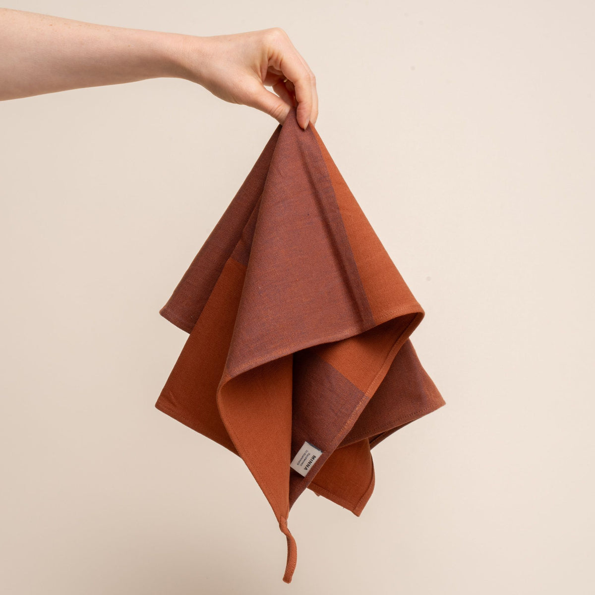 A hand holding a tea towel with large color blocks of rust and dark red, and featuring a hanging loop on a corner