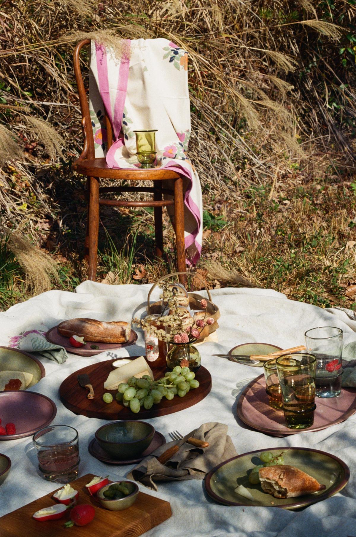 An outdoor picnic with plates and food on a white blanket with a wooden chair and tall grass in the background