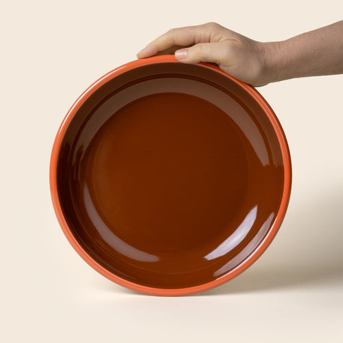 A hand props up an enamel dinner plate in dark red-brown with an orange rim.