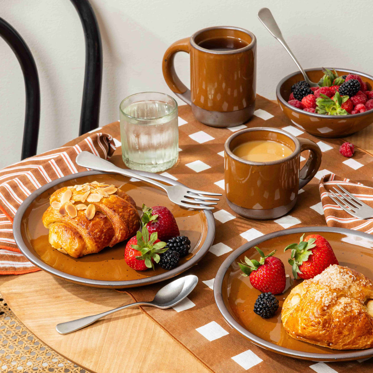 A cozy breakfast scene featuring two brown ceramic plates with pastries topped with almonds and sugar, alongside fresh strawberries and blackberries. Two matching mugs filled with coffee and a bowl of mixed berries are set on a wooden table with a checkered tablecloth and striped napkins.
