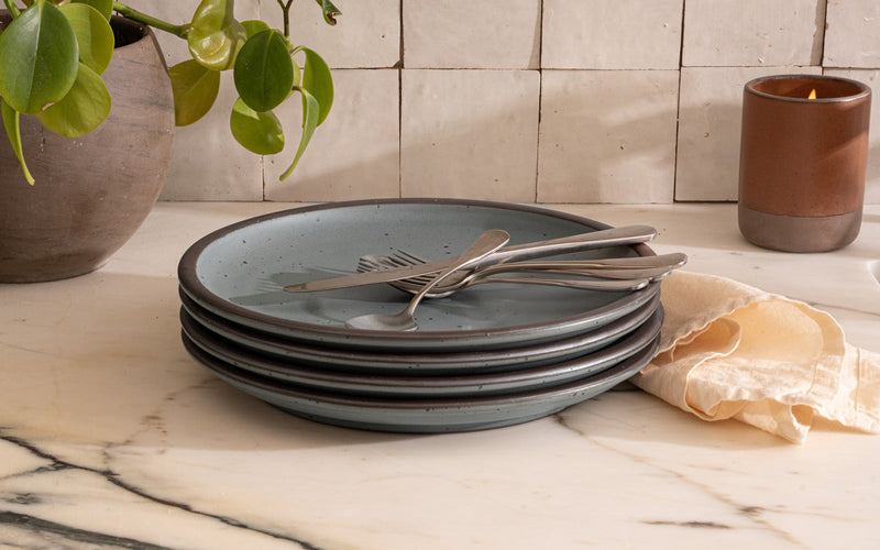 A stack of 4 dinner sized ceramic plates in a blue-grey color featuring iron speckles and an unglazed rim, with silverware and napkin on a countertop