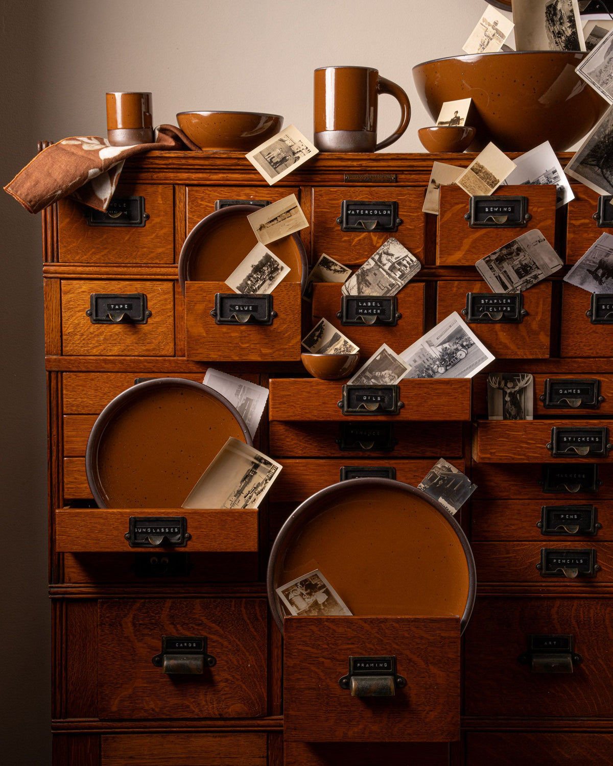 A wooden cabinet with multiple drawers, some partially open, contains caramel-colored ceramic dishes, a mug, and vintage photographs cascading out. A folded cloth rests on the cabinet, complementing the warm hues of the ceramics.