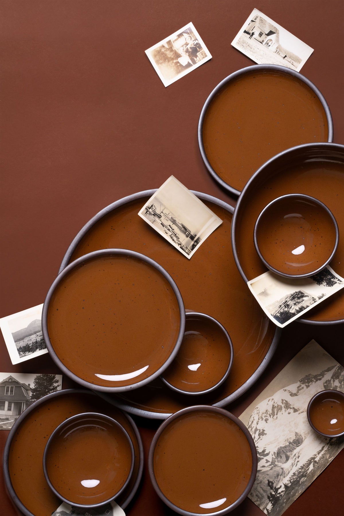 A collection of brown glazed ceramic plates and bowls arranged on a warm brown background, interspersed with vintage black-and-white photographs.