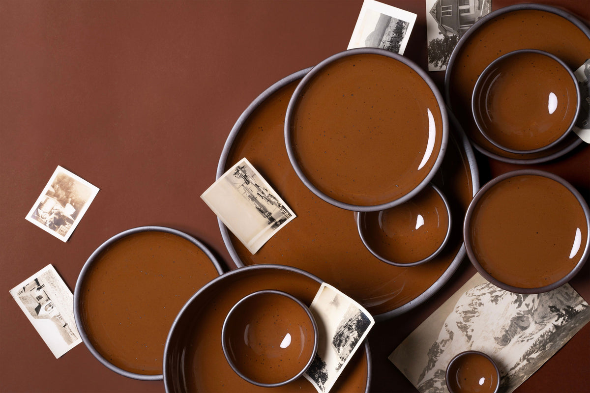 Assortment of brown glazed ceramic plates and bowls from East Fork, arranged with vintage black-and-white photographs on a matching brown background.