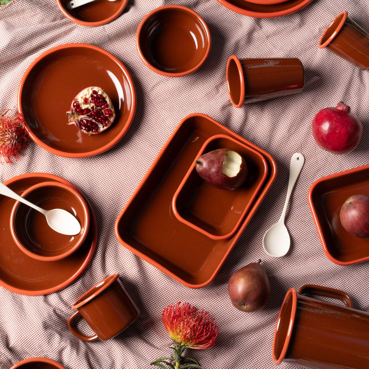 Top-down view of glossy rust-colored enamel dishes scattered with pears, pomegranates, spoons, and flowers on a textured cloth.