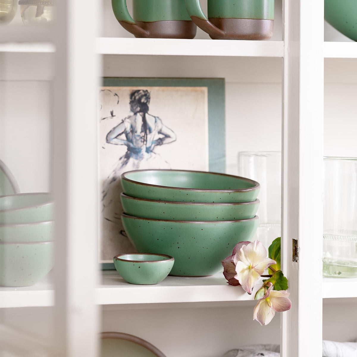 Stacked green ceramic bowls on a white shelf beside a framed sketch and a small flower, viewed through a cabinet frame.