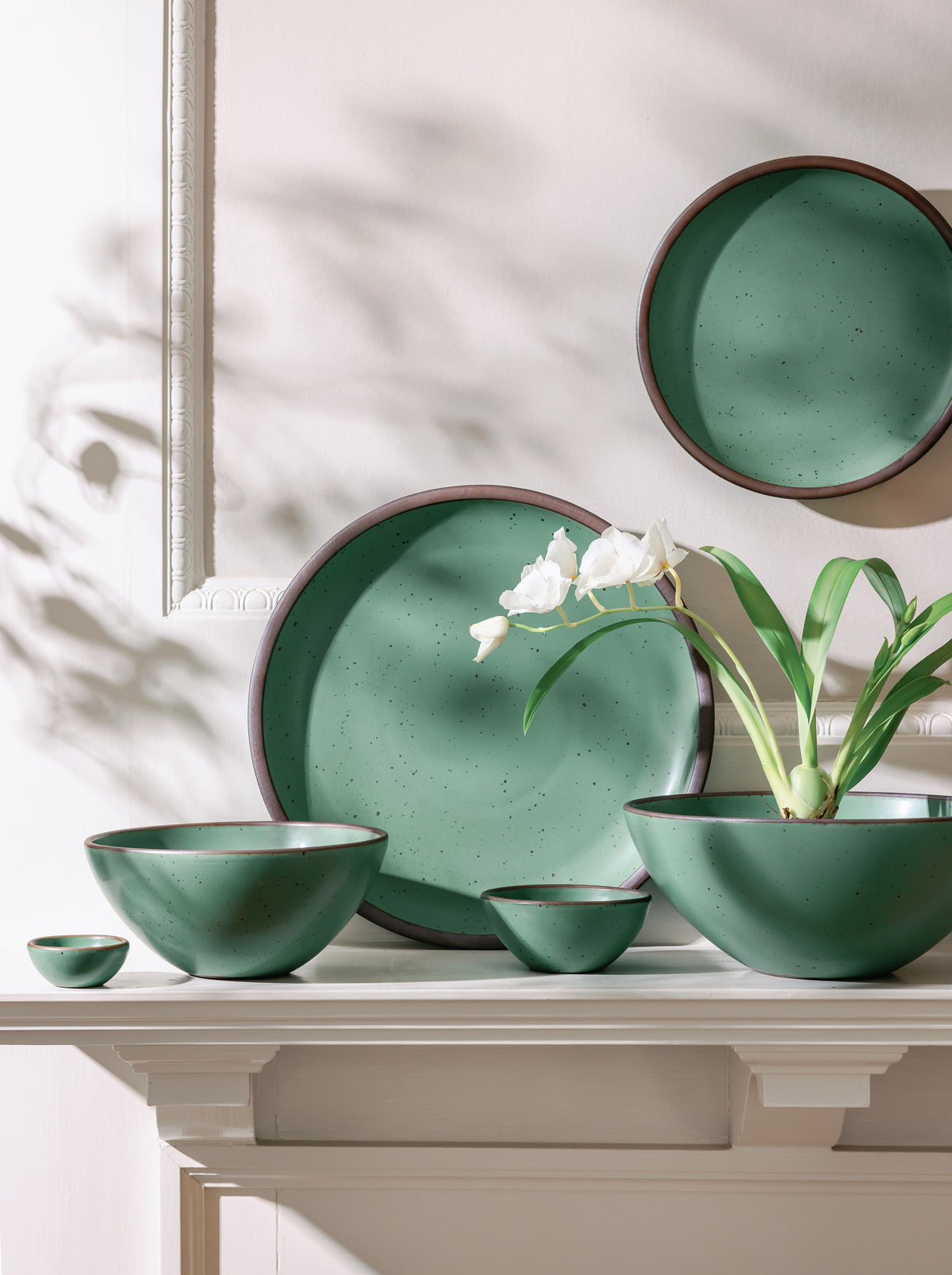 A set of green ceramic bowls and a plate arranged on a mantel, with a white flowering plant placed in one of the bowls, lit by soft natural light.