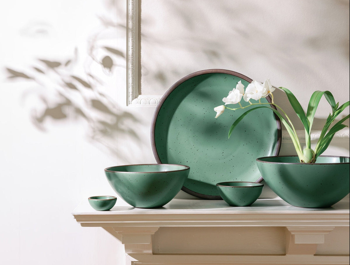 A set of green ceramic bowls and a plate arranged on a mantel, with a white flowering plant placed in one of the bowls, lit by soft natural light.