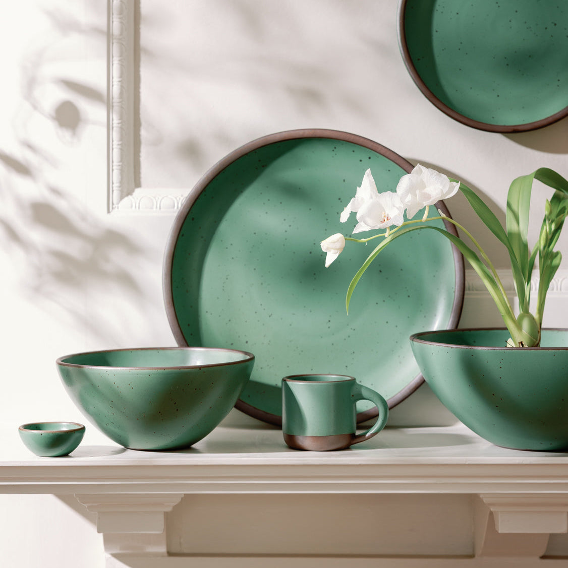Green ceramic tableware set on a white mantle surface with a plant and flower decoration.