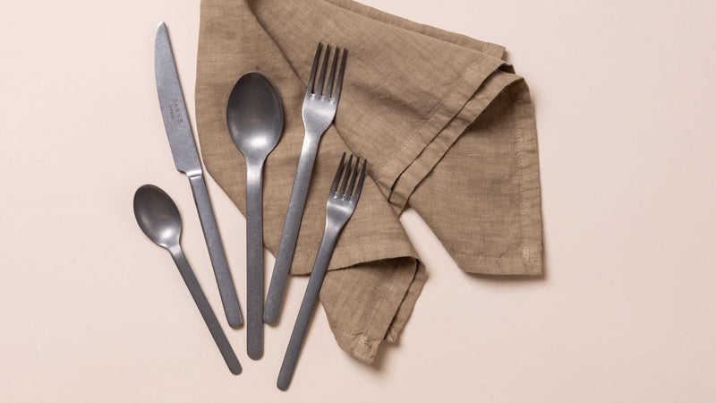 Set of silver cutlery including knives, forks, and spoons on a beige napkin against a beige background.