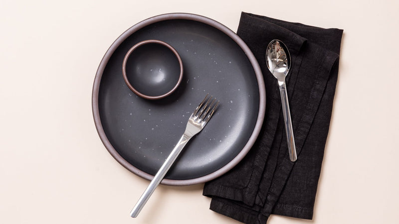 An overhead view of ceramic plates in graphite balck with iron speckles, and a matching napkin with a steel flatware.