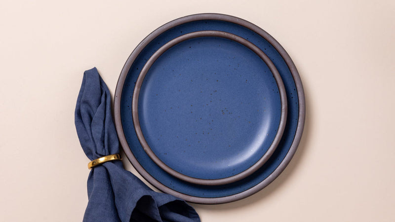 An overhead view of ceramic plates in blue with iron speckles, and a matching napkin with a brass napkin ring.