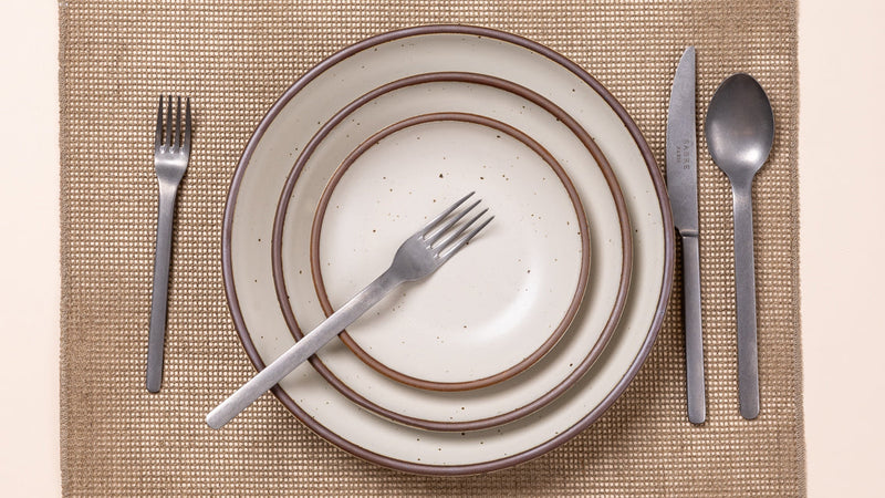 An overhead view of ceramic plates in a warm off-white, with steel flatware and woven natural placemat.